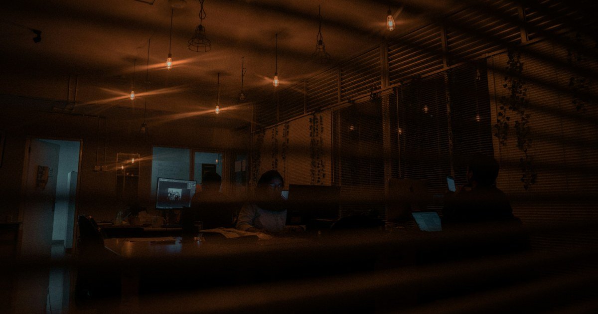 Escritório pequeno no fim do dia, com uma pessoa ainda sentada à mesa sob a luz de uma luminária